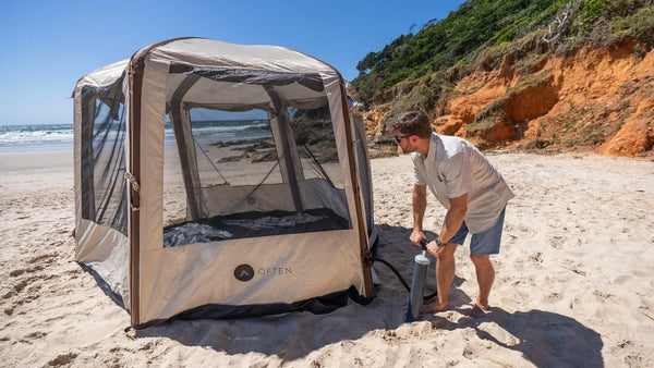 A camper inflates an Often Bunker hexagonal tent with a hand pump on an Australian beach, the tent already standing with no poles and an ochre headland behind.