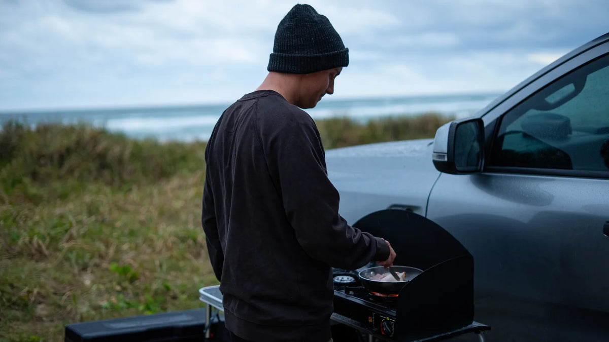 A camper cooks in a frying pan on a two-burner portable gas stove mounted on a folding kitchen at the tailgate of a 4WD ute, with coastal dune grass and breaking surf behind.