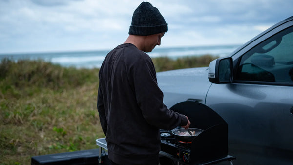 A camper cooks in a frying pan on a two-burner portable gas stove mounted on a folding kitchen at the tailgate of a 4WD ute, with coastal dune grass and breaking surf behind.