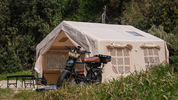 Often canvas tent seen on the diagonal from a low angle, with a Dirodi electric bike parked in front and the camp table beside it, in coastal scrub