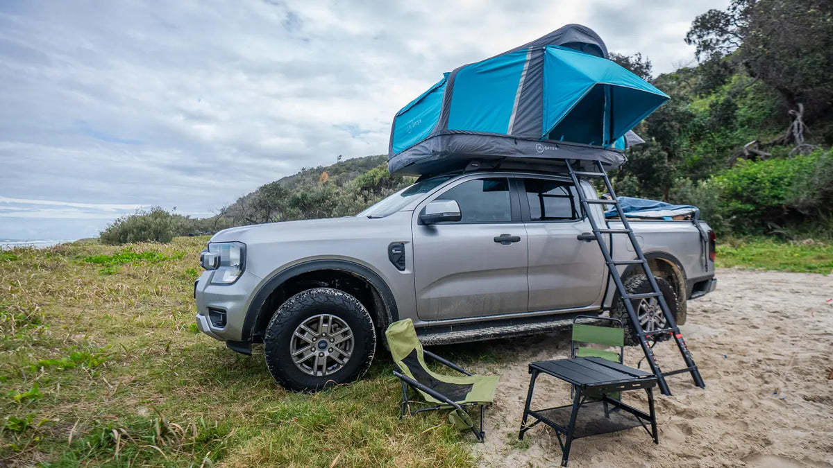 Ford Ranger with an Often inflatable rooftop tent open and the ladder down, camp chairs and a low table set up beside it on a grassy dune, with the surf behind