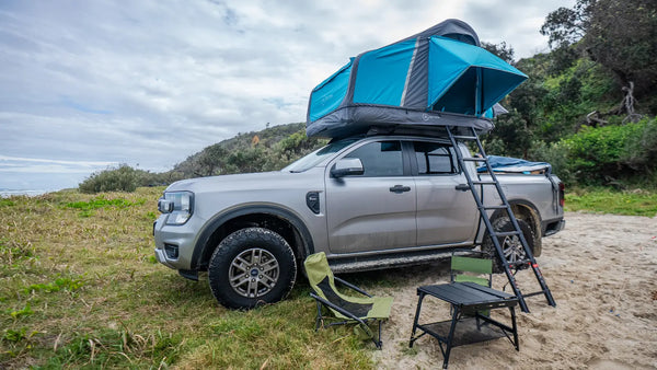 Ford Ranger with an Often inflatable rooftop tent open and the ladder down, camp chairs and a low table set up beside it on a grassy dune, with the surf behind