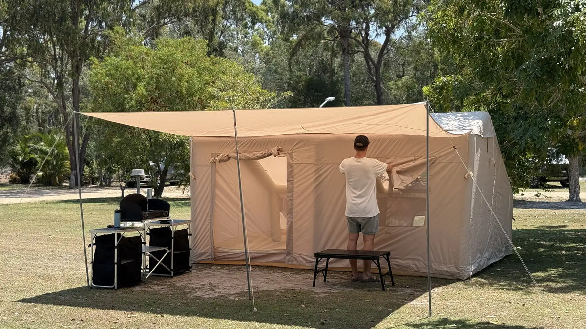 A camper adjusts the wall of a large beige canvas tent with its awning extended at a grassy Queensland campground, a folding camp kitchen set up beside it and gum trees behind.