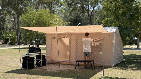 A camper adjusts the wall of a large beige canvas tent with its awning extended at a grassy Queensland campground, a folding camp kitchen set up beside it and gum trees behind.