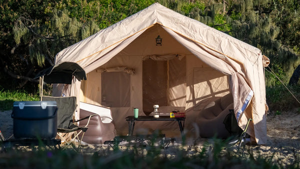 A ground tent open at first light on a grassy campsite with the sun low behind the treeline and a chair and mug outside the door.
