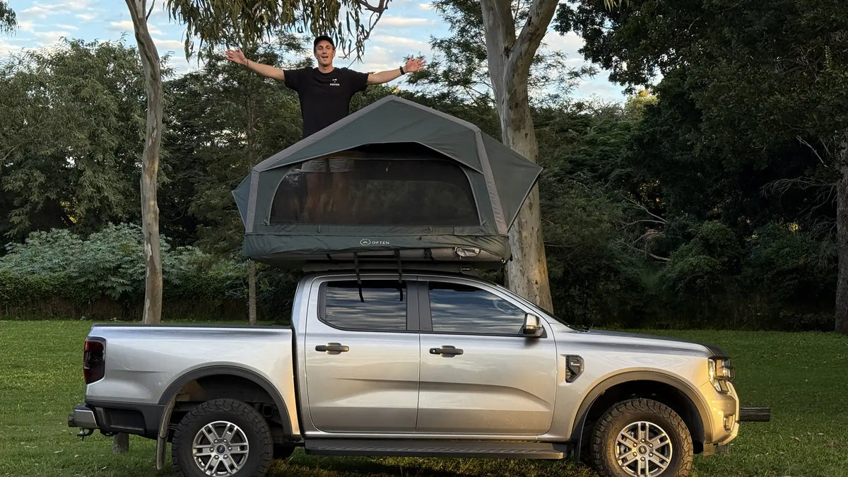 A silver 4WD ute parked side-on at a grassy campground with an Often inflatable rooftop tent open on its roof rack, and a person standing in the tray beside the tent with their arms out, gum trees behind.