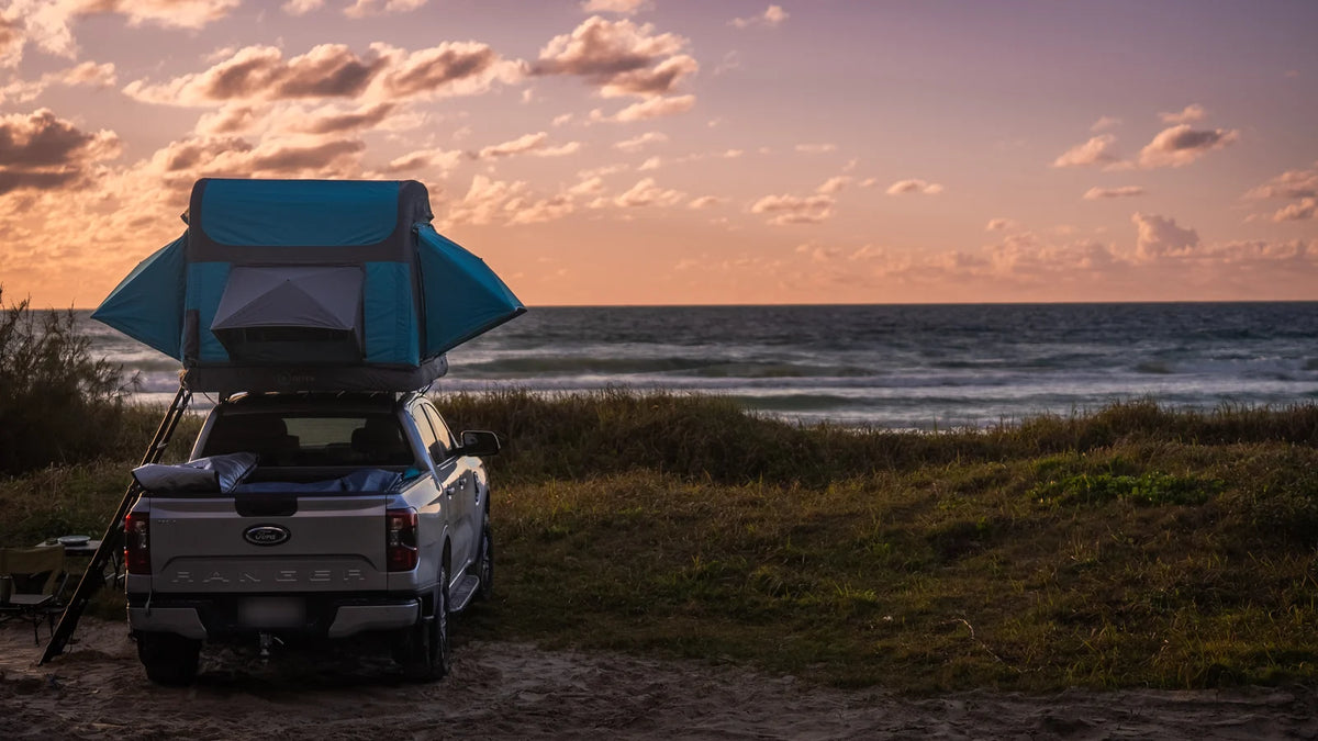 An Often rooftop tent open on a 4WD ute parked on a coastal dune at last light, the sea behind and the camp set up for the night, with dune grass in the foreground.
