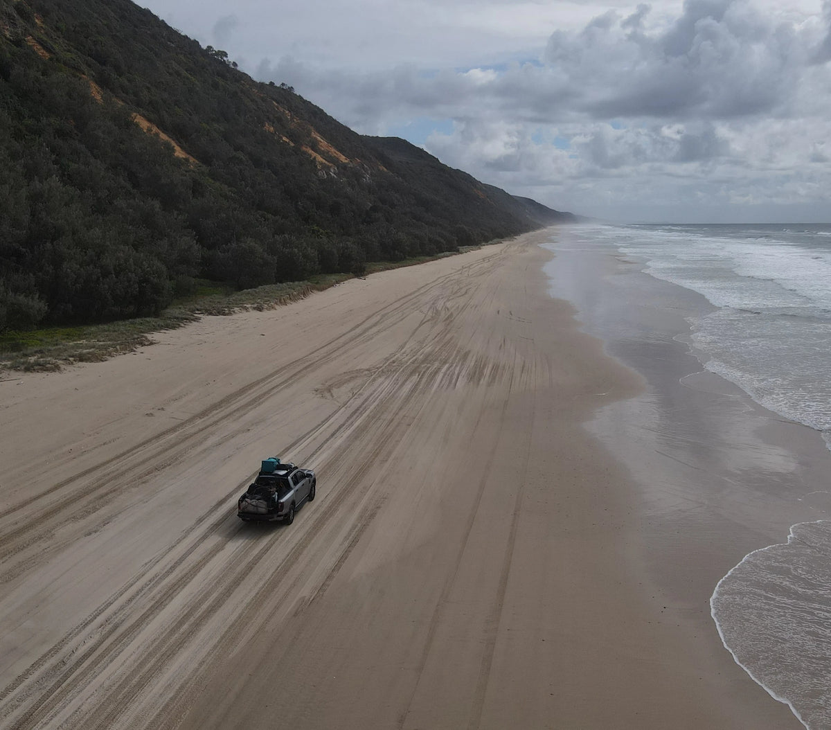 A loaded four wheel drive driving alone along a wide firm-sand beach on the Queensland coast, seen from a drone, with the forested headland receding into the distance