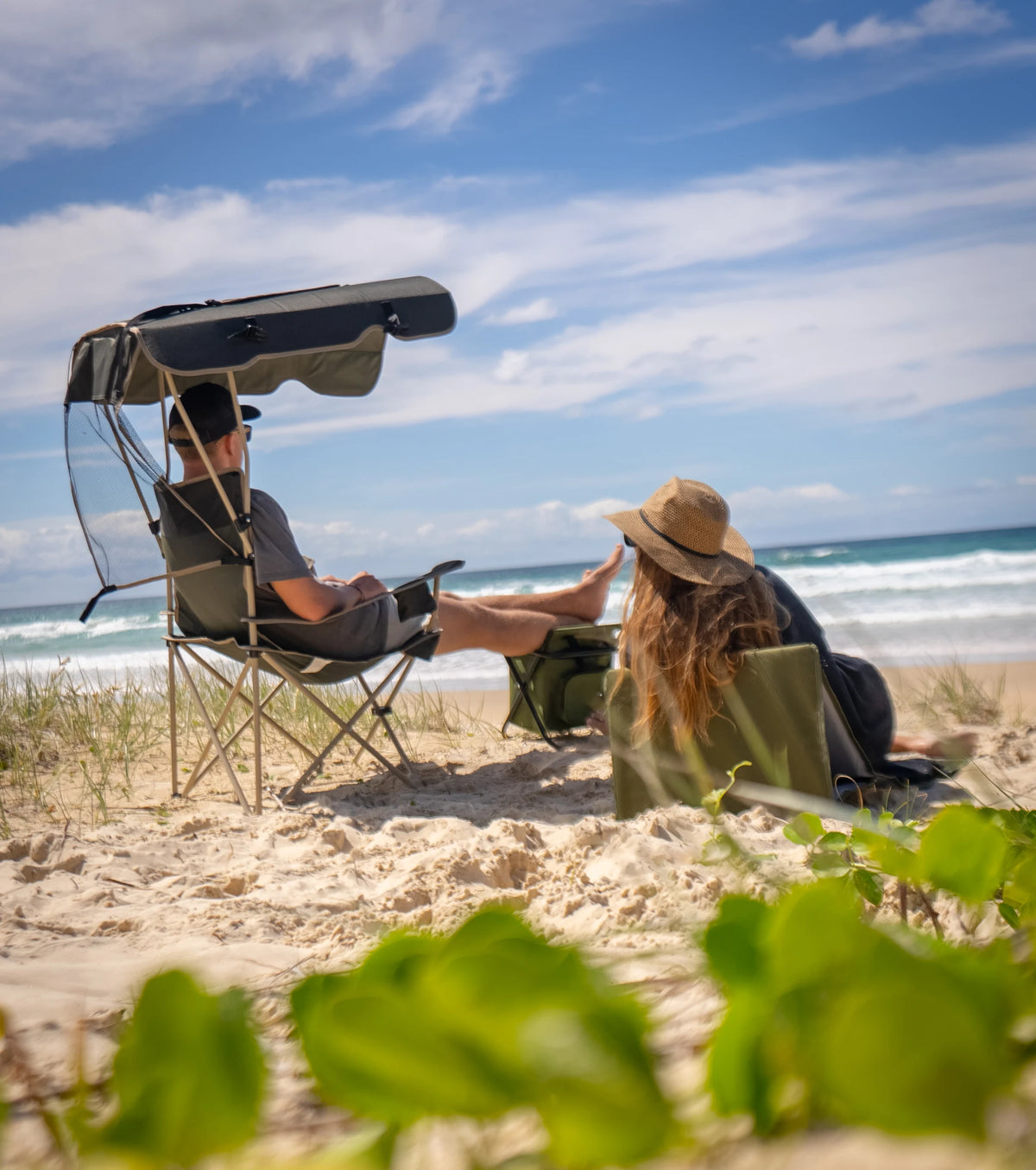 A couple on a Queensland beach: a man reclining in an Often camping chair with its own sun canopy, and a woman in a low chair beside him, both looking out at the surf.