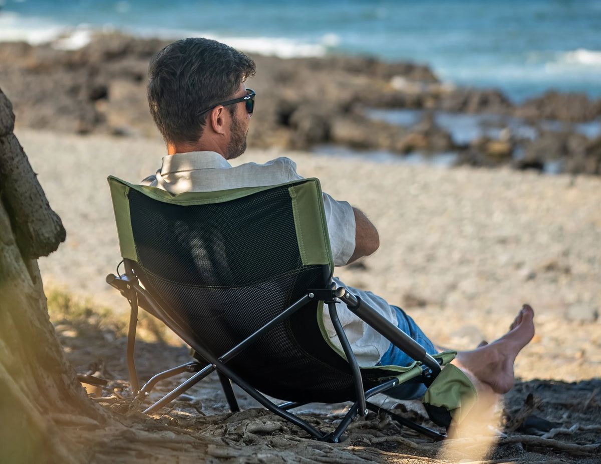 A man sitting in an Often low mesh-back camping chair in the shade of a palm, on the sand of a rocky stretch of the Queensland coast.