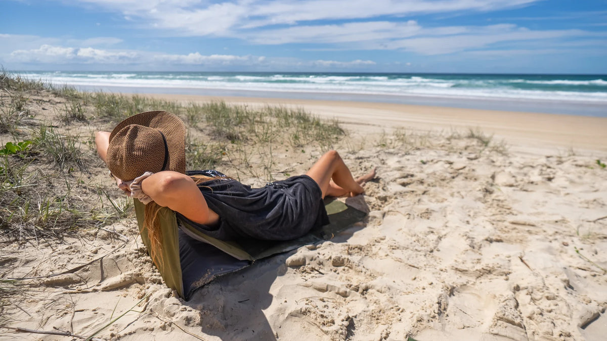 A woman lying back on an Often canvas deck chair laid flat on a sand dune, hat over her face