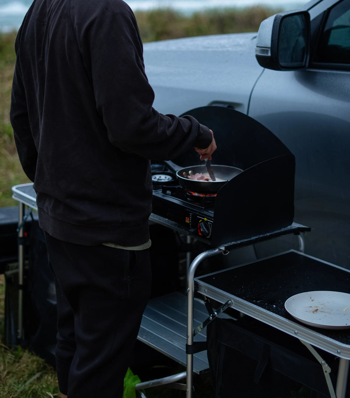 A man cooking at a camp kitchen set up at the back of a Ford Ranger, with the grey sea behind him.
