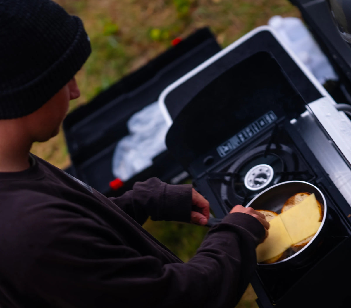 A man in a beanie tipping food out of a pan while cooking outdoors at first light, at the back of a ute.