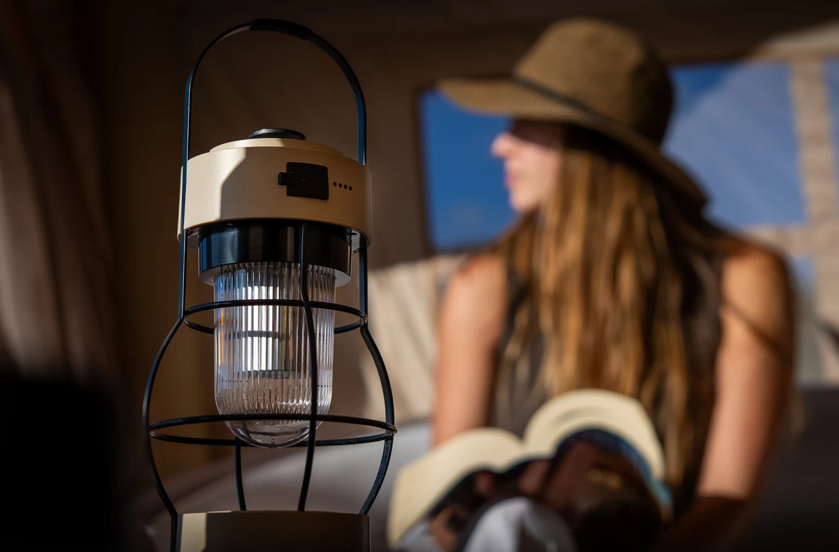An Often retro camping lantern standing in the foreground inside a glamping tent, with a woman reading a book on the inflatable bed behind it.